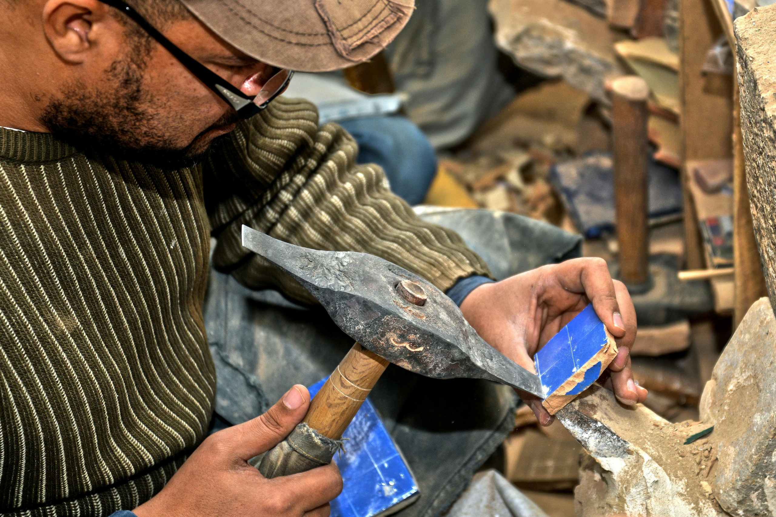 Close-up of a craftsman sharpening a blade on a stone, wearing a striped sweater and cap, with wooden tool handles nearby.